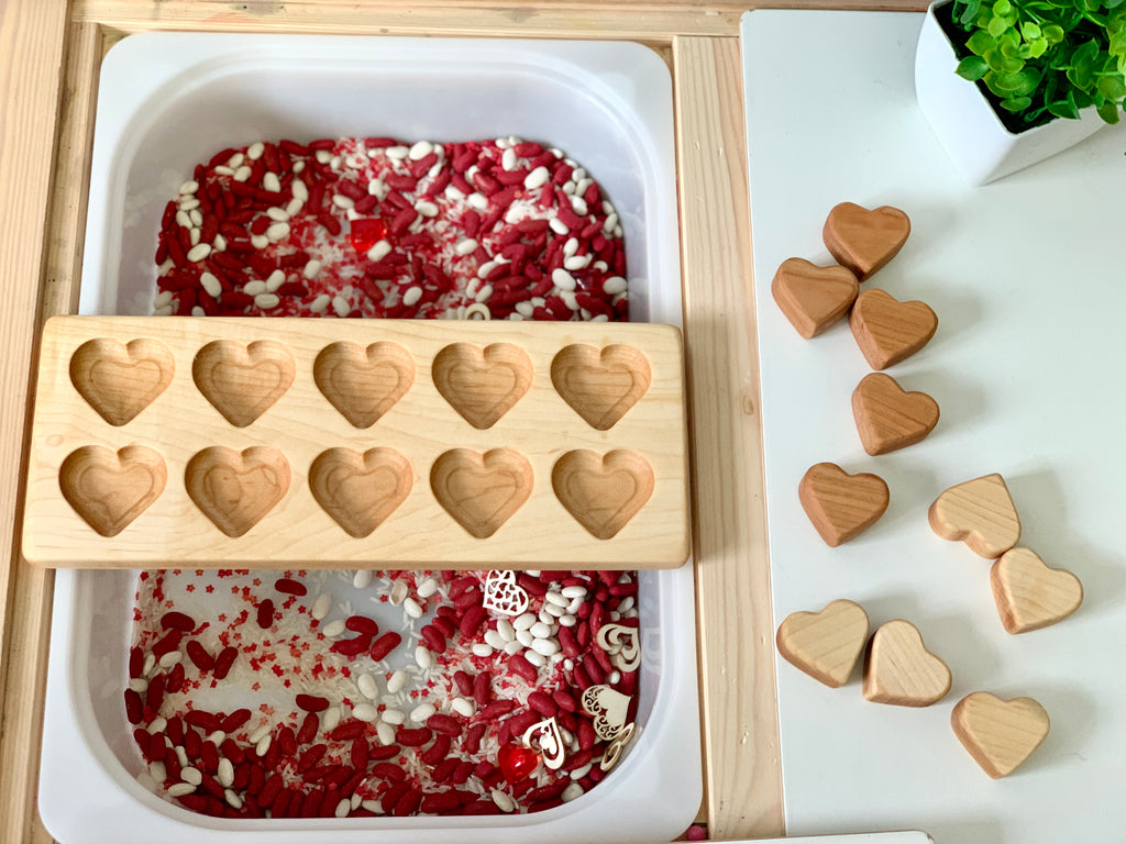 Wooden heart-shaped molds and solid wooden hearts next to a white bin filled with red and white sensory filler beads