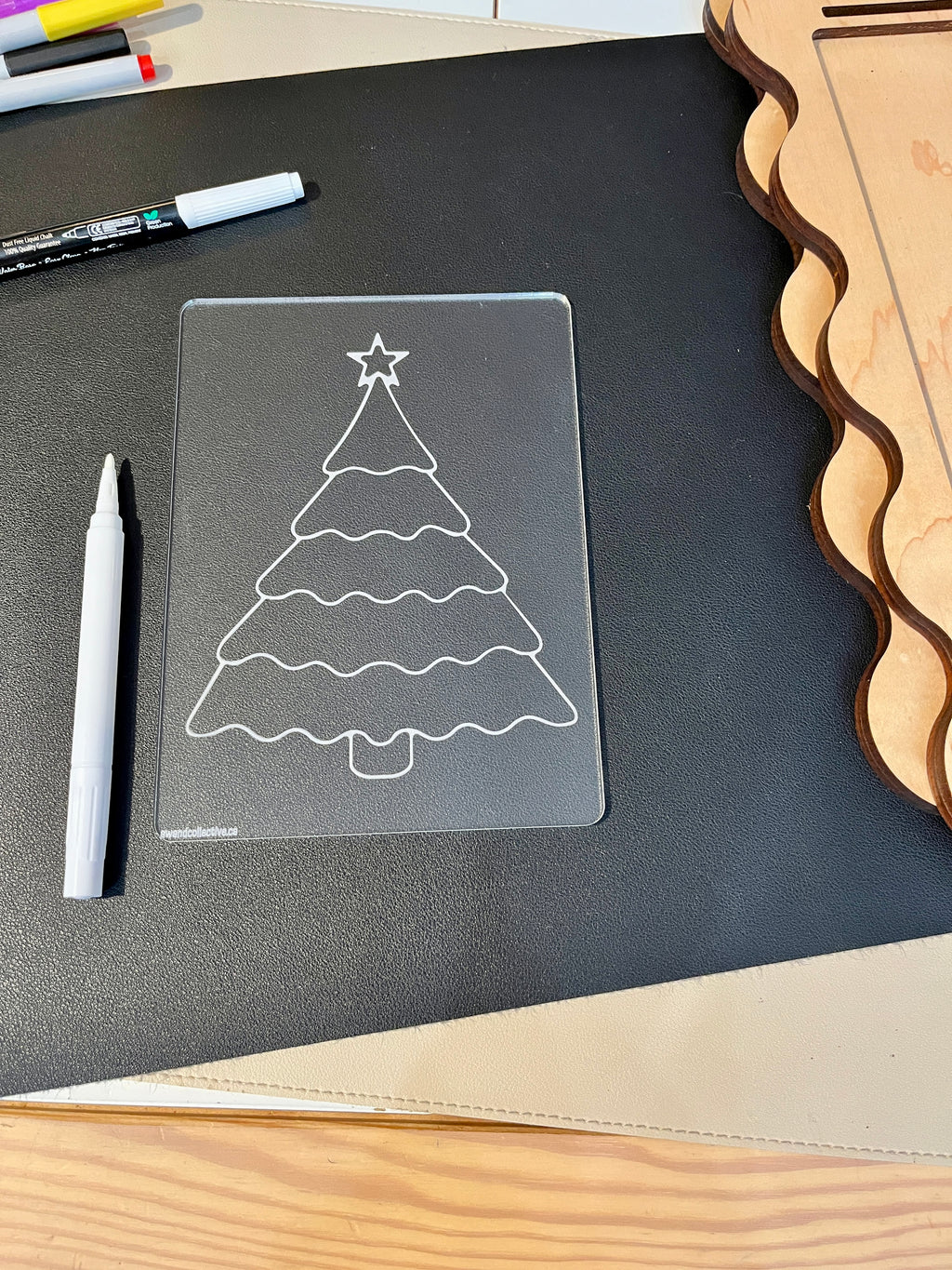 Transparent plastic board with outlined Christmas tree shape and star on a black surface beside markers and wooden cutouts