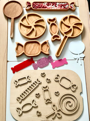 Wooden trays and puzzle pieces shaped like candies and lollipops with red and white beans and colorful paper cutouts on white surface