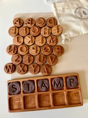 Round wooden alphabet discs with recessed uppercase letters arranged beside a wooden tray holding letter-shaped clay pieces and a fabric drawstring bag