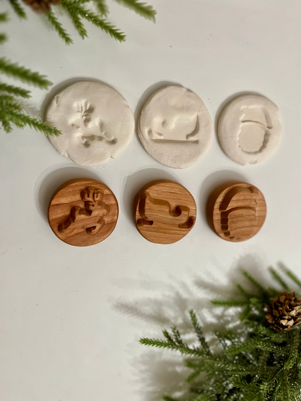Three wooden holiday stamps with reindeer, sleigh, and Santa hat shapes alongside matching white clay imprints on white surface with pine branches