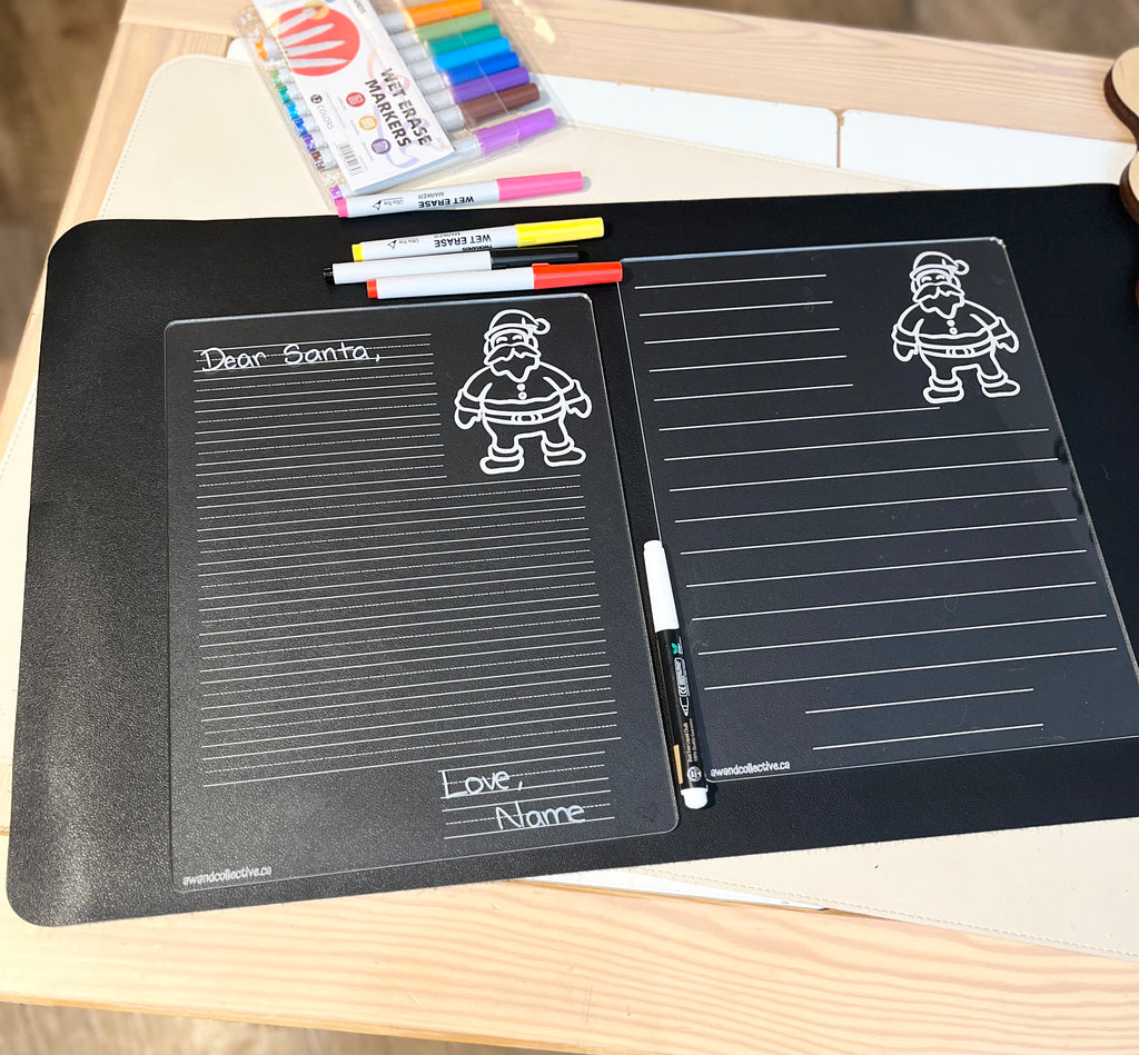 Clear acrylic writable board with lined sections and Santa Claus illustration, set on black surface with colorful markers nearby