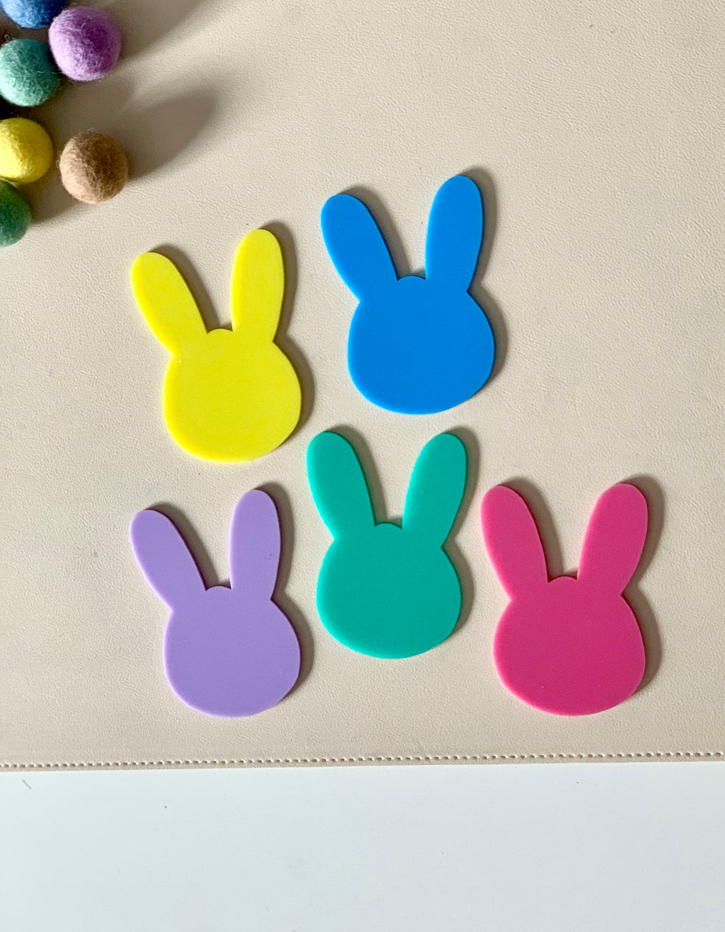 Five colorful bunny-shaped cutouts in yellow, blue, purple, teal, and pink on a beige surface near felt balls