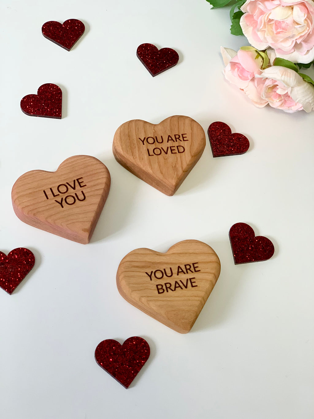 Three wooden heart-shaped blocks engraved with "I LOVE YOU," "YOU ARE LOVED," and "YOU ARE BRAVE" surrounded by red glitter hearts