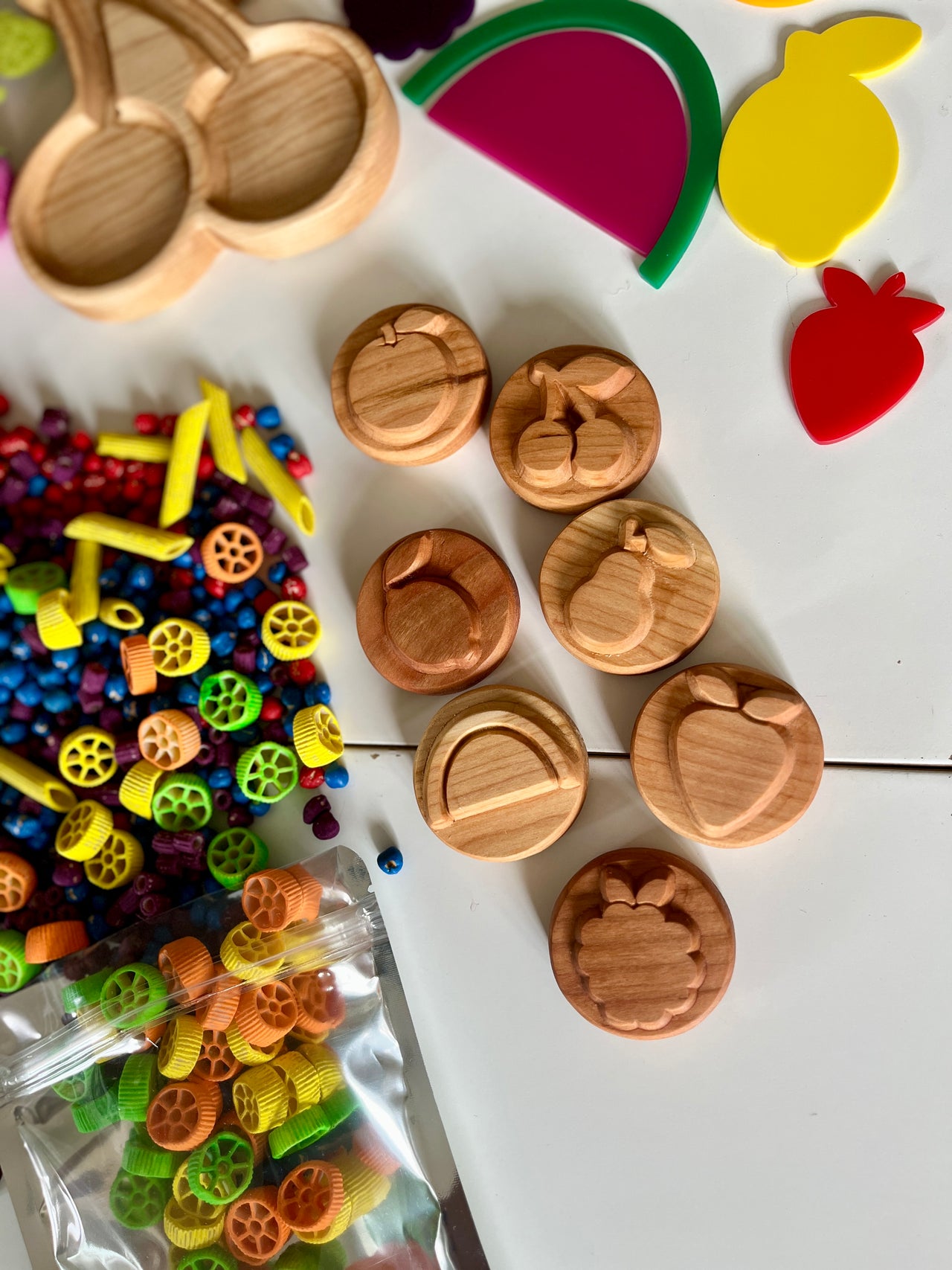 Seven round wooden fruit stamps with colorful pasta and fruit-shaped play trays on white surface