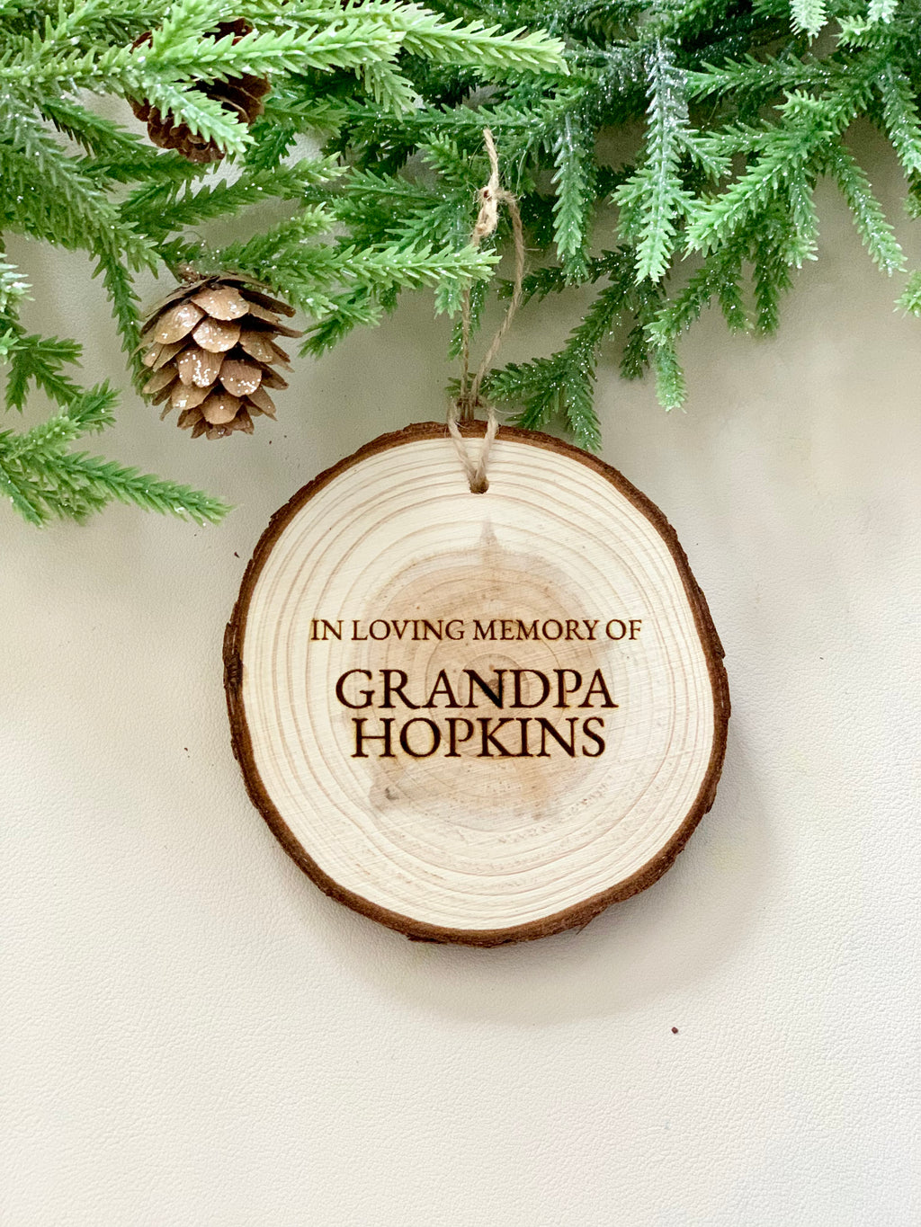 Round wooden memorial Christmas ornament engraved with "In Loving Memory of Grandpa Hopkins" hanging from twine under pine branches