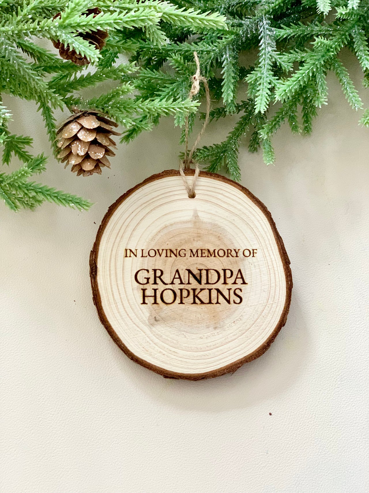 Round wooden memorial Christmas ornament engraved with "In Loving Memory of Grandpa Hopkins" hanging from twine under pine branches