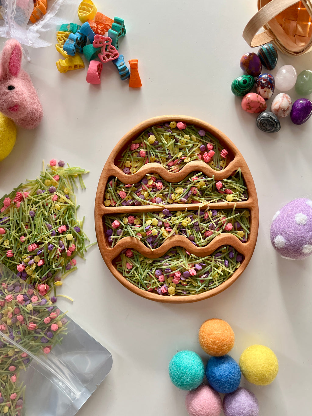 Round wooden tray filled with green pasta and small colorful flower-shaped sprinkles with felt craft decorations nearby