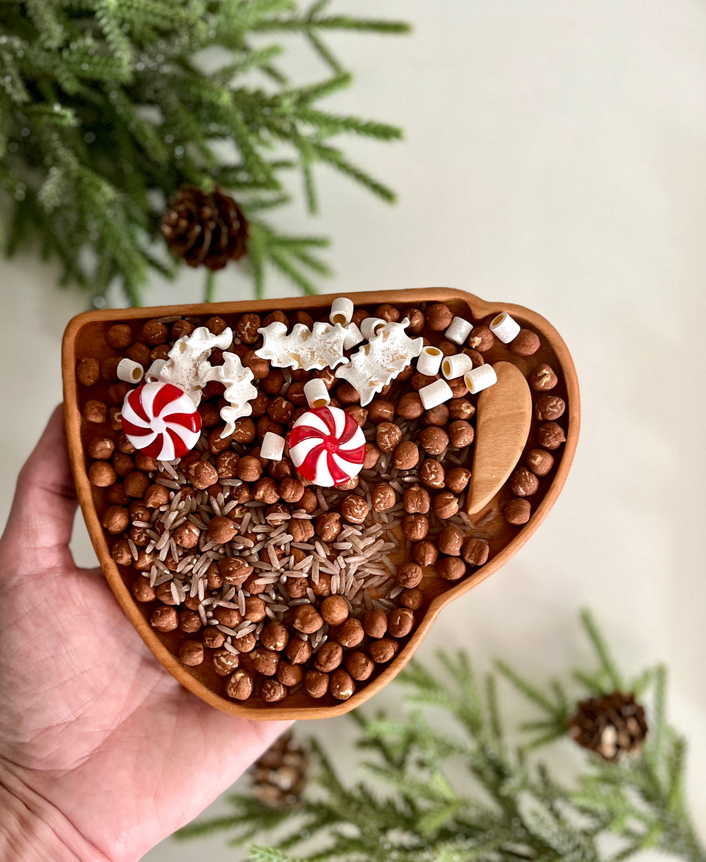 Hand holding wooden cup-shaped tray filled with hazelnuts, rice, white candy pieces, red peppermint candies, and wooden scoop