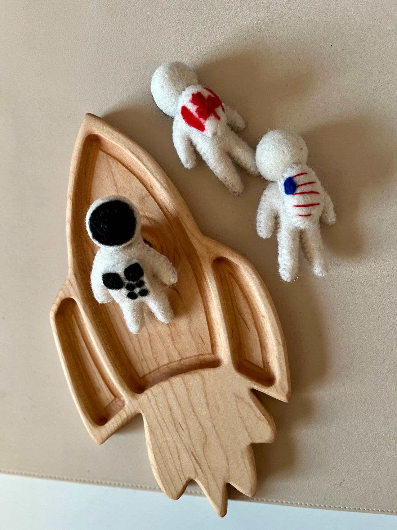 Three small handmade felt astronaut figures with black, red, and blue details placed on and around a wooden rocket tray on a beige surface