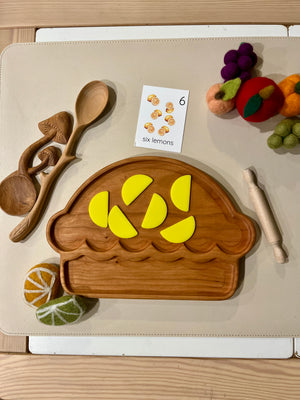 Wooden muffin-shaped tray with six bright yellow lemon wedge pieces, wooden utensils, felt fruit, and a counting card nearby