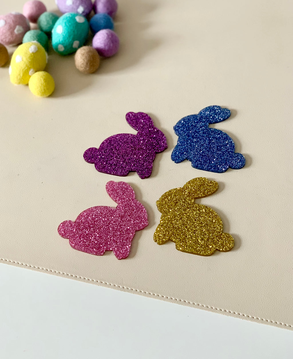 Four glitter-covered bunny cutouts in purple, blue, pink, and gold on a beige surface near pastel felt balls