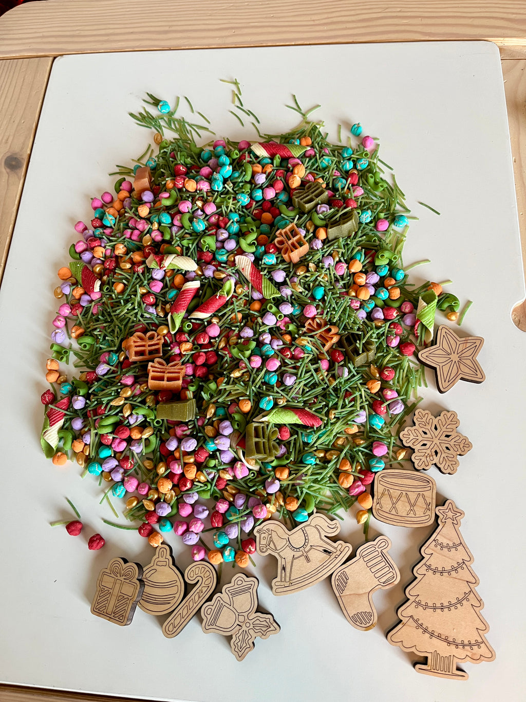 Colorful mix of small beads, green dried pine needles, striped pasta, and wooden Christmas-themed cutouts on white board