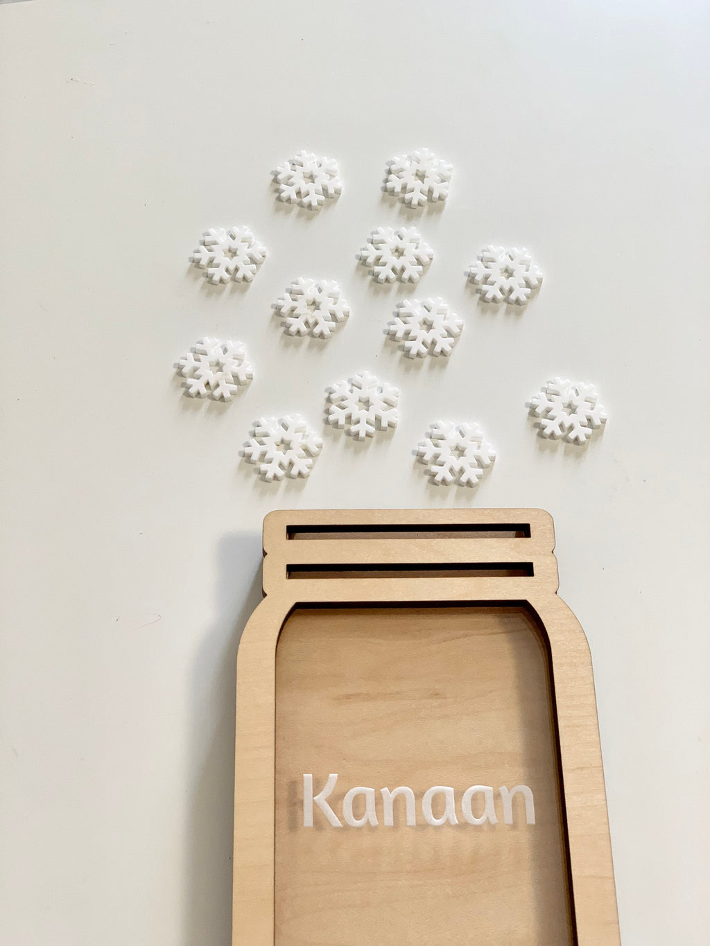 Light wooden jar-shaped frame with the name Kanaan and scattered white snowflake tokens on a light surface