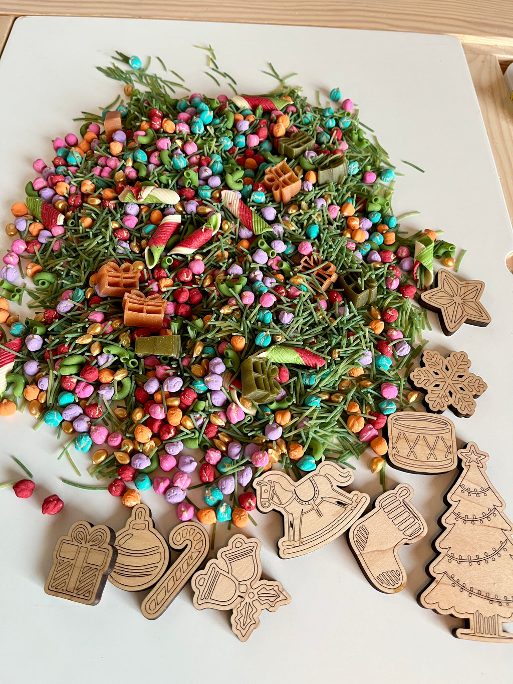 Colorful sensory mix of beads, dried botanicals, pasta, and wooden Christmas-themed cutouts on white surface