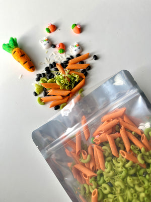 Clear bag spilling orange and green pasta with black beans, white rice, and small bunny and carrot decorations on a white surface