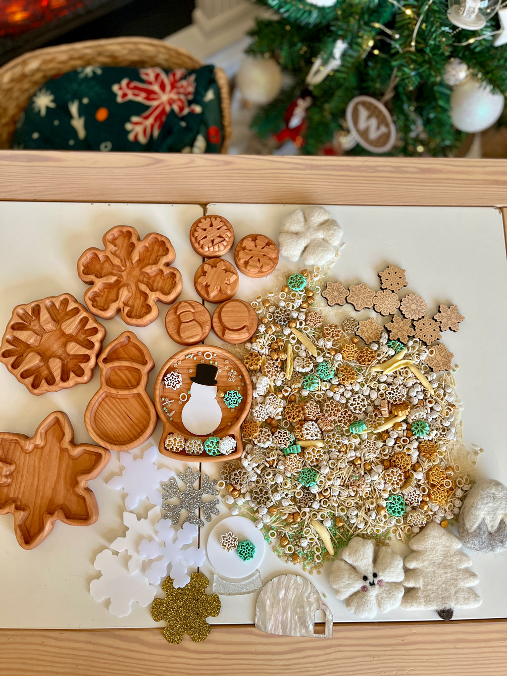 Wooden snowman and snowflake-shaped trays alongside natural and craft sensory materials on white tabletop near decorated Christmas tree