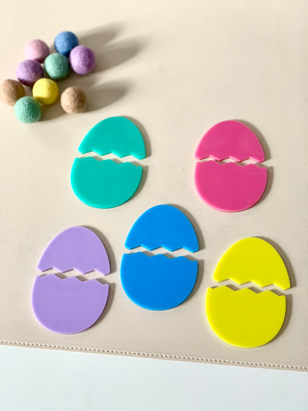 Five pastel-colored broken egg-shaped cutouts in turquoise, pink, purple, blue, and yellow with felt balls nearby on a beige surface