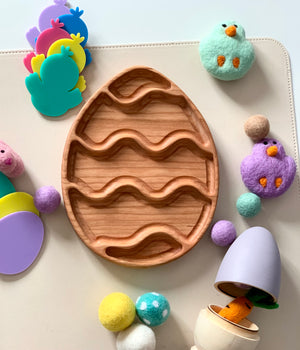 Wooden egg-shaped tray with carved wavy grooves surrounded by felt balls, plush chicks, colorful chick cutouts, and a toy carrot in a wooden egg container