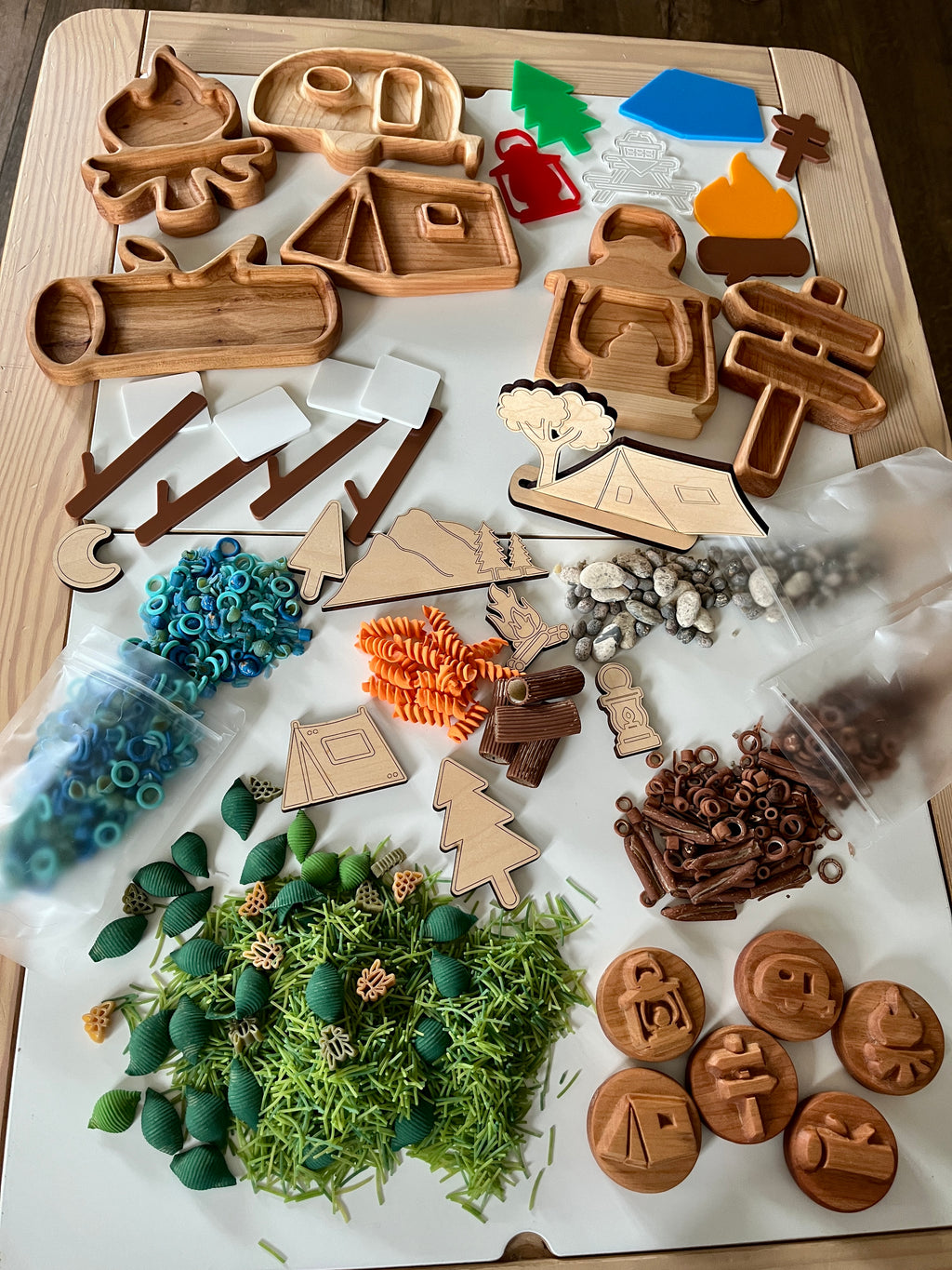 Wooden camping-themed shapes and tokens with colorful plastic rings, pasta, pebbles, and shredded natural materials on white surface