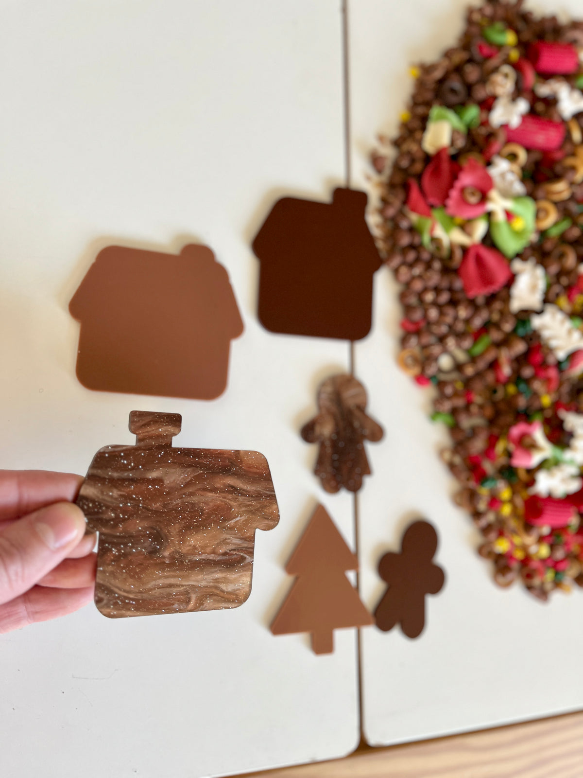 Hand holding brown marbled house-shaped cutout with other brown holiday shapes and colorful sensory materials nearby