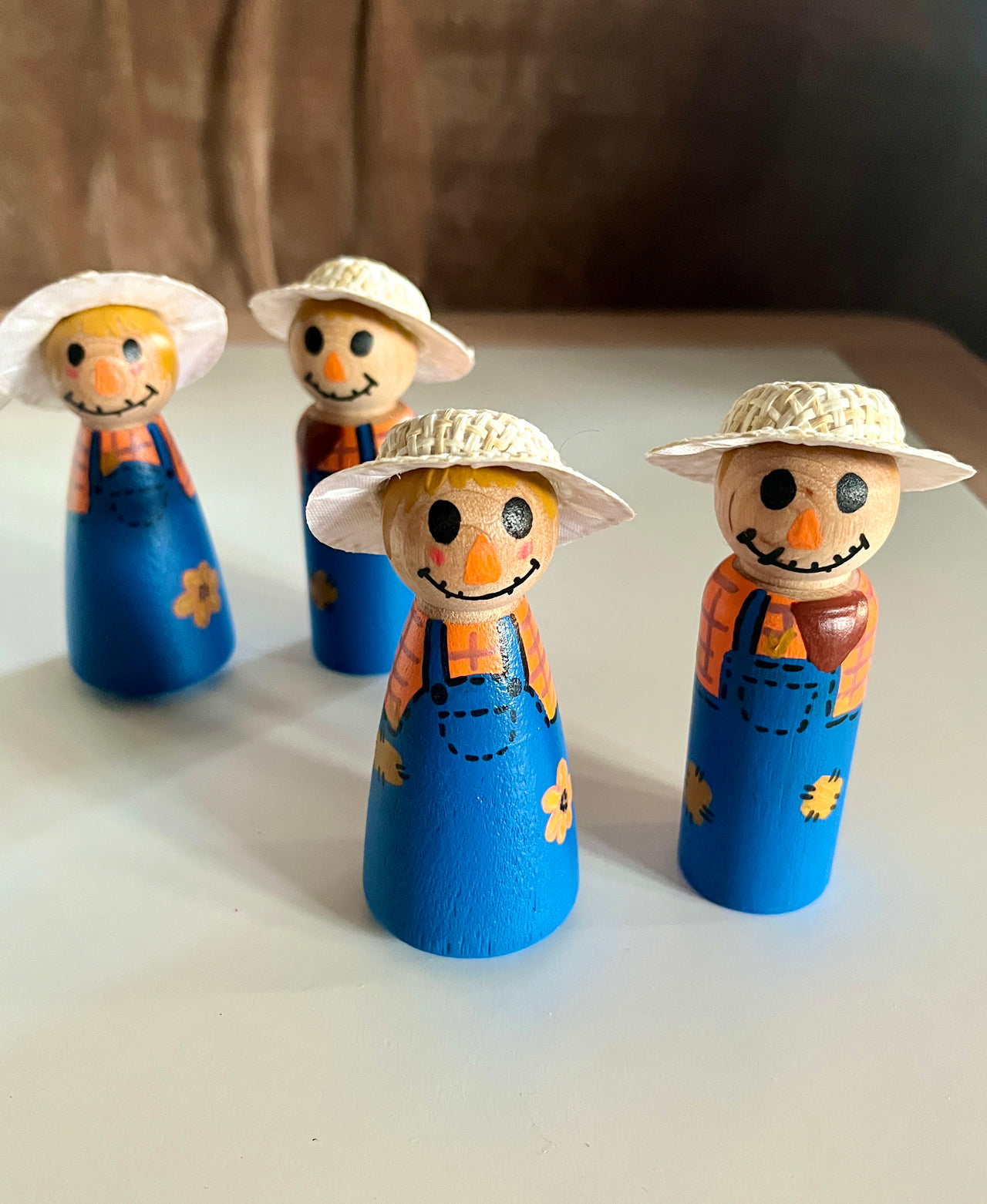 Four wooden scarecrow figurines wearing blue overalls and straw hats arranged on a white surface