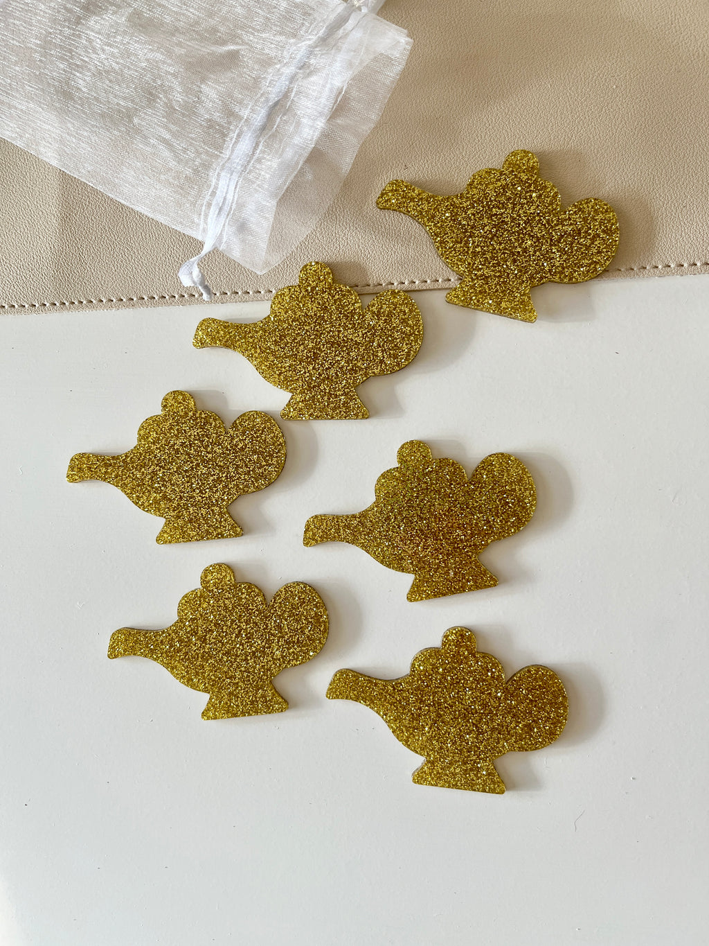 Six gold glitter genie lamp-shaped pieces arranged near a white sheer drawstring pouch on a two-tone surface