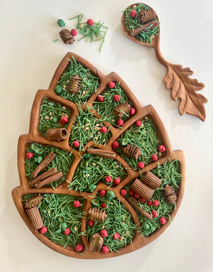Wooden leaf-shaped tray and matching spoon holding green herbs and small red berries on white background