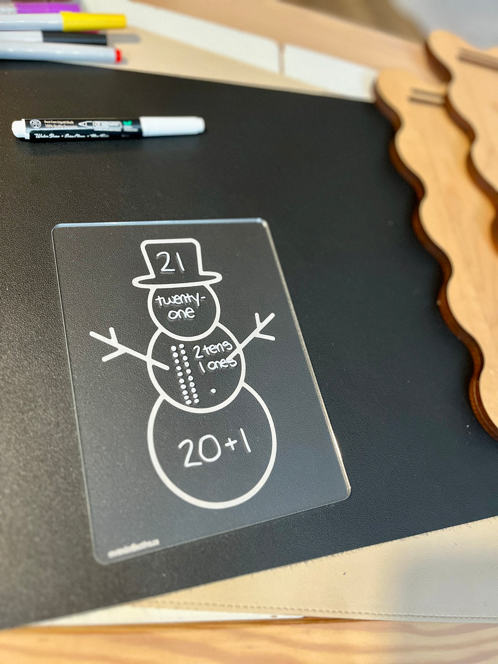 Clear plastic board with a snowman diagram illustrating the number 21 as tens and ones breakdown on a black surface