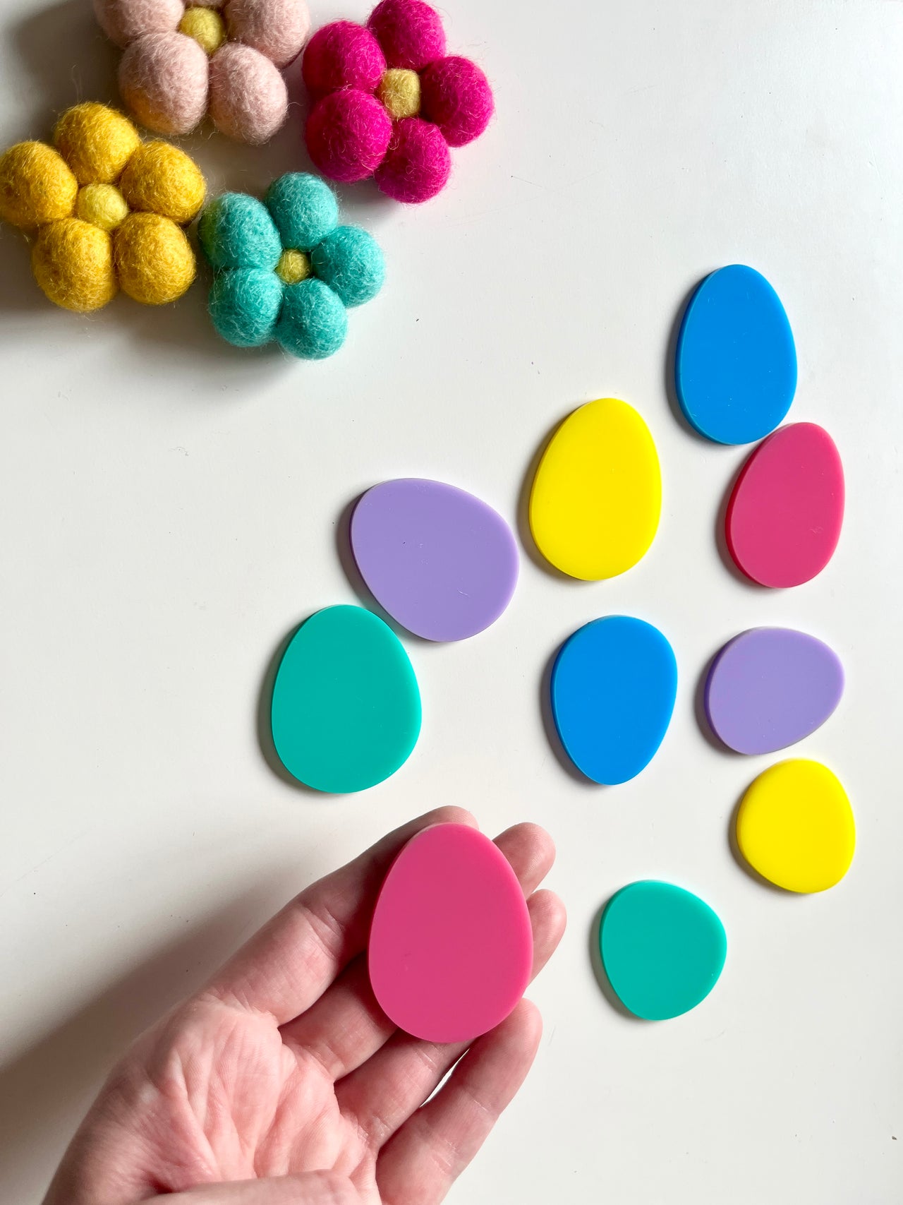 Hand holding pink egg-shaped plastic piece surrounded by scattered colorful pastel egg shapes and felt flowers on white surface