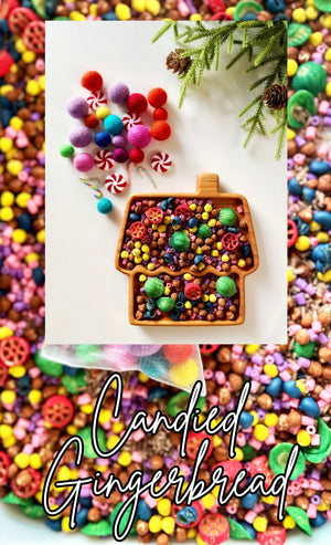 Wooden tray shaped like a gingerbread house filled with colorful beads and surrounded by pom-poms and greenery on white surface