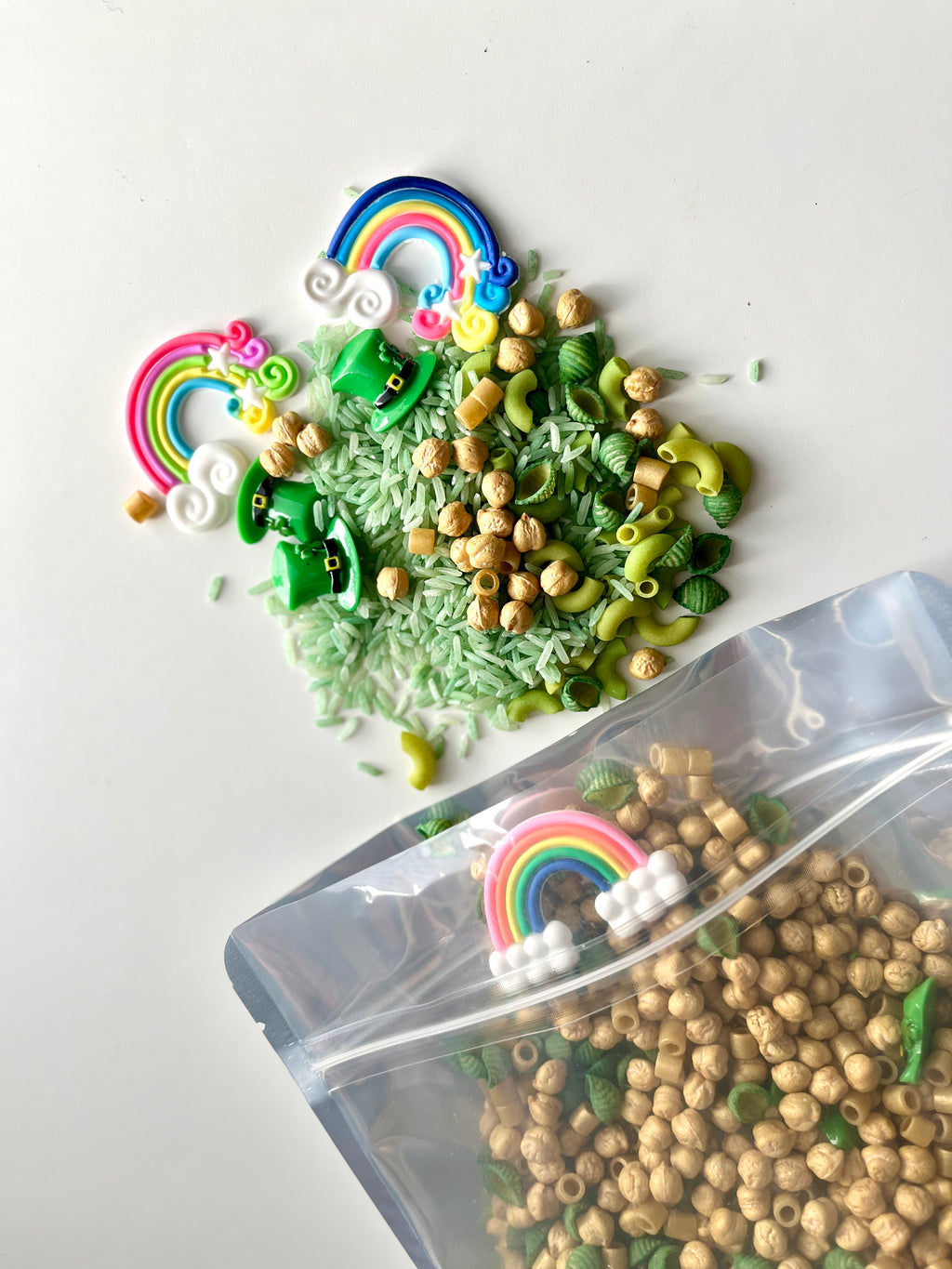 Clear plastic bag spilling natural beads, green pasta, dyed rice, green shamrocks, leprechaun hats, and rainbow decorations on white surface