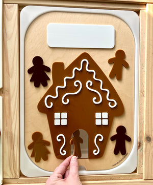 Child's hand placing a brown gingerbread figure into a decorated gingerbread house cutout on a wooden activity board