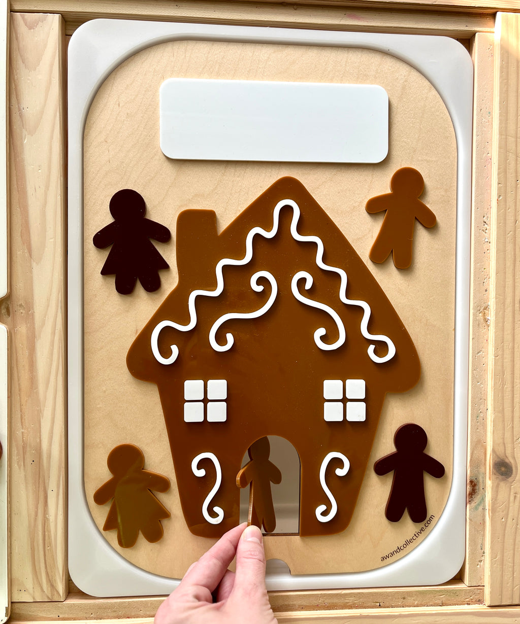 Child's hand placing a brown gingerbread figure into a decorated gingerbread house cutout on a wooden activity board