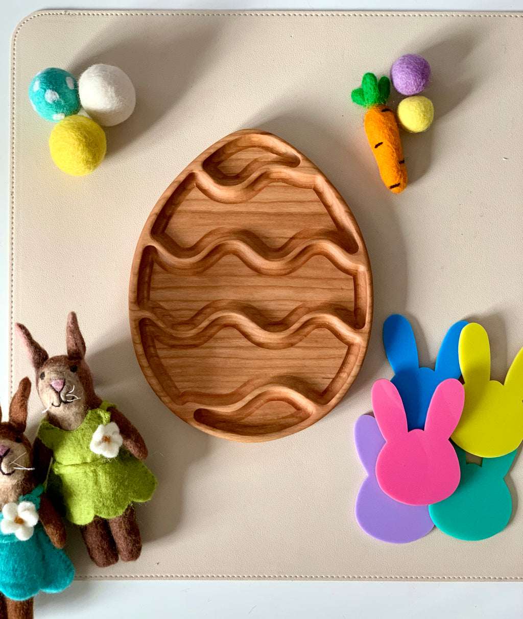 Wooden egg-shaped tray with carved wave patterns surrounded by felt balls, two plush rabbits, felt carrot, and colorful bunny cutouts