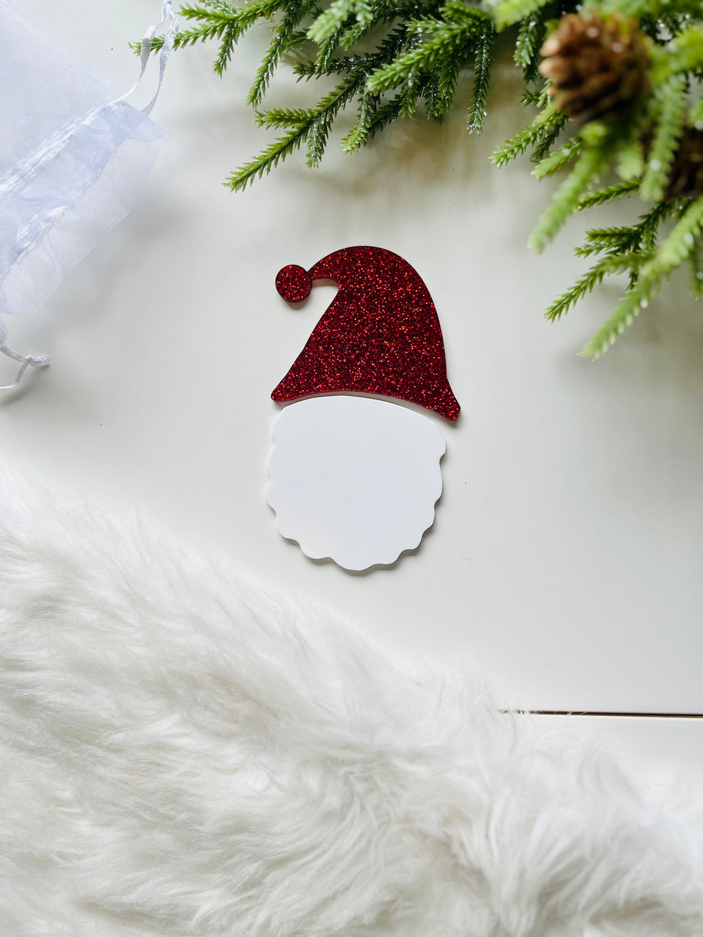 Flat red glitter Santa hat and white beard cutout on white surface with greenery and white fur accents nearby
