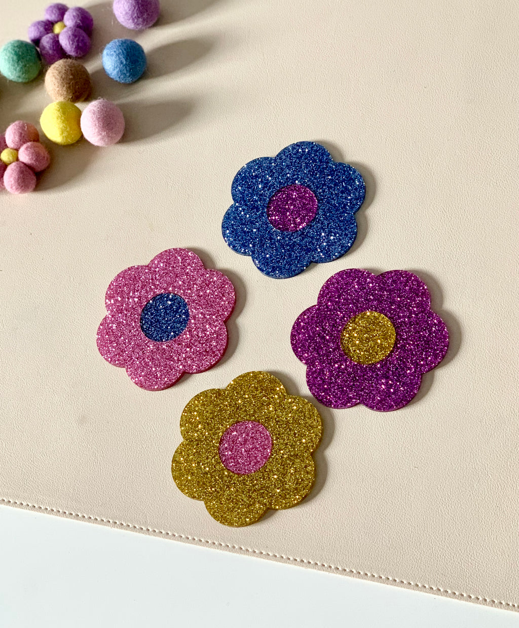 Four glittery flower-shaped cutouts in gold, pink, purple, and blue on a beige surface near colorful felt balls