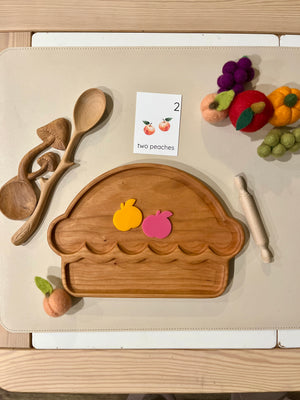 Wooden pie-shaped tray with two colorful peach-shaped cutouts, wooden utensils, and felt fruit on beige surface