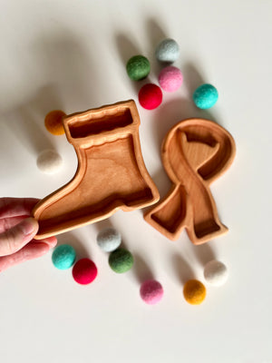 Hand holding wooden tray shaped like a stocking next to a ribbon-shaped tray surrounded by colorful felt balls on white surface