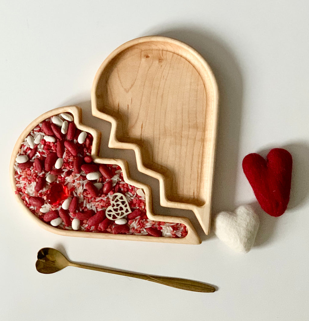Heart-shaped wooden tray with one half filled with red and white beans and a small carved heart, next to felt hearts and a gold spoon