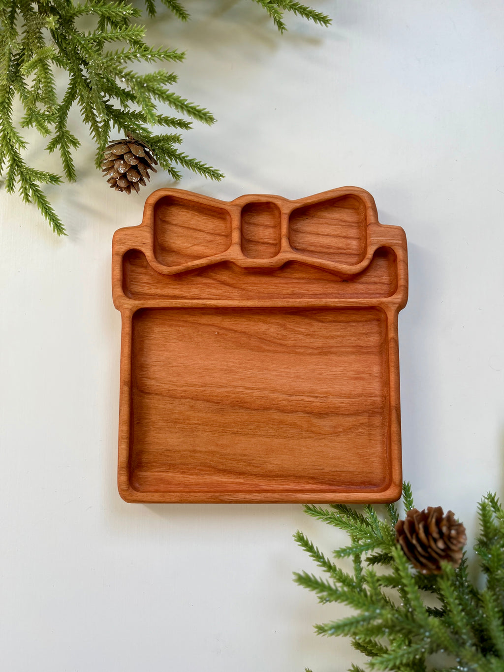 Handcrafted wooden tray shaped like a gift box with bow, surrounded by pine branches and pine cones on white surface