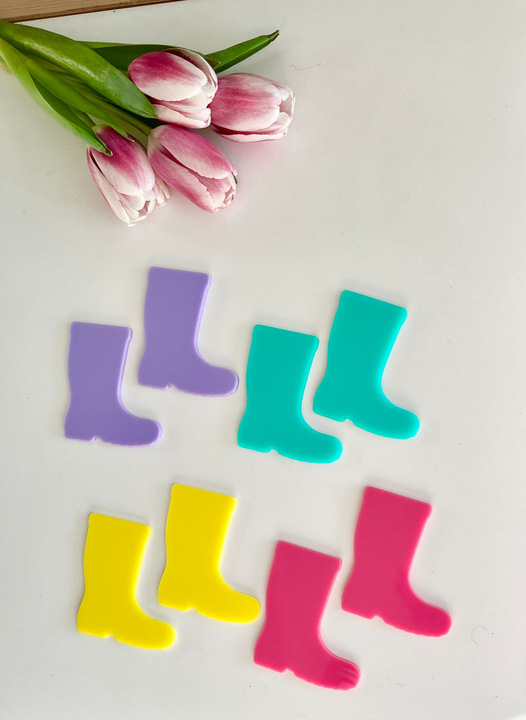 Four pairs of colorful rain boot cutouts in purple, blue, yellow, and pink beside pink and white tulips on white surface