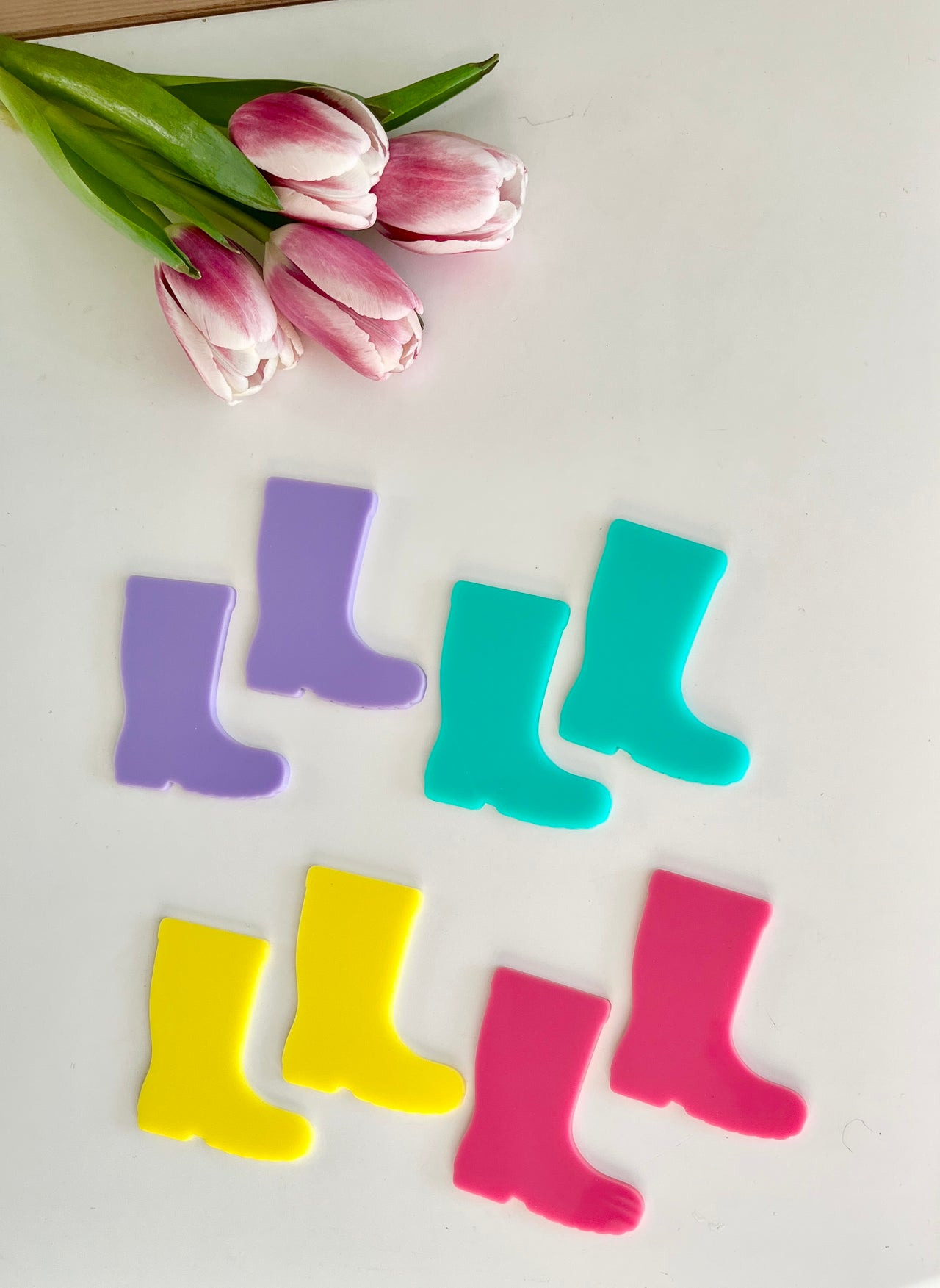 Four pairs of colorful rain boot cutouts in purple, blue, yellow, and pink beside pink and white tulips on white surface