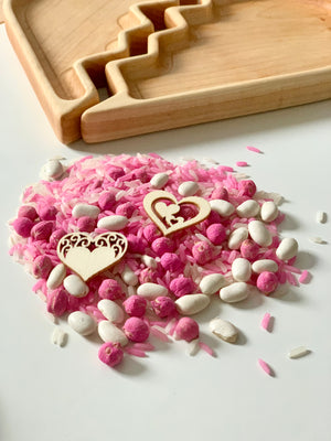 Pink and white sensory rice with small white beans and two wooden hearts next to a wooden tray with heart cutouts