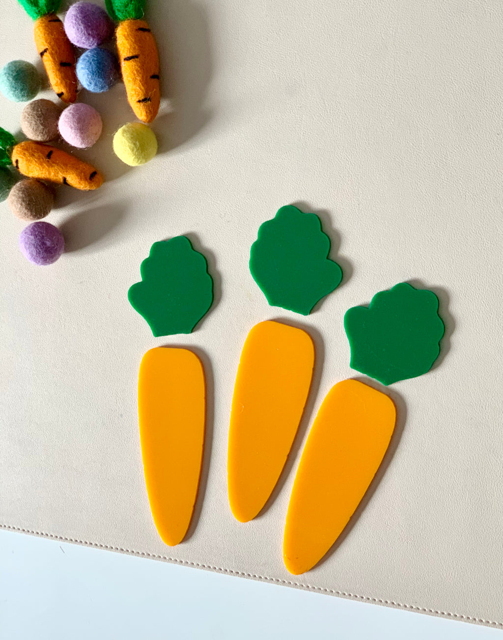 Three flat orange carrot-shaped cutouts with green tops on a light surface near colorful felt balls and fabric carrots