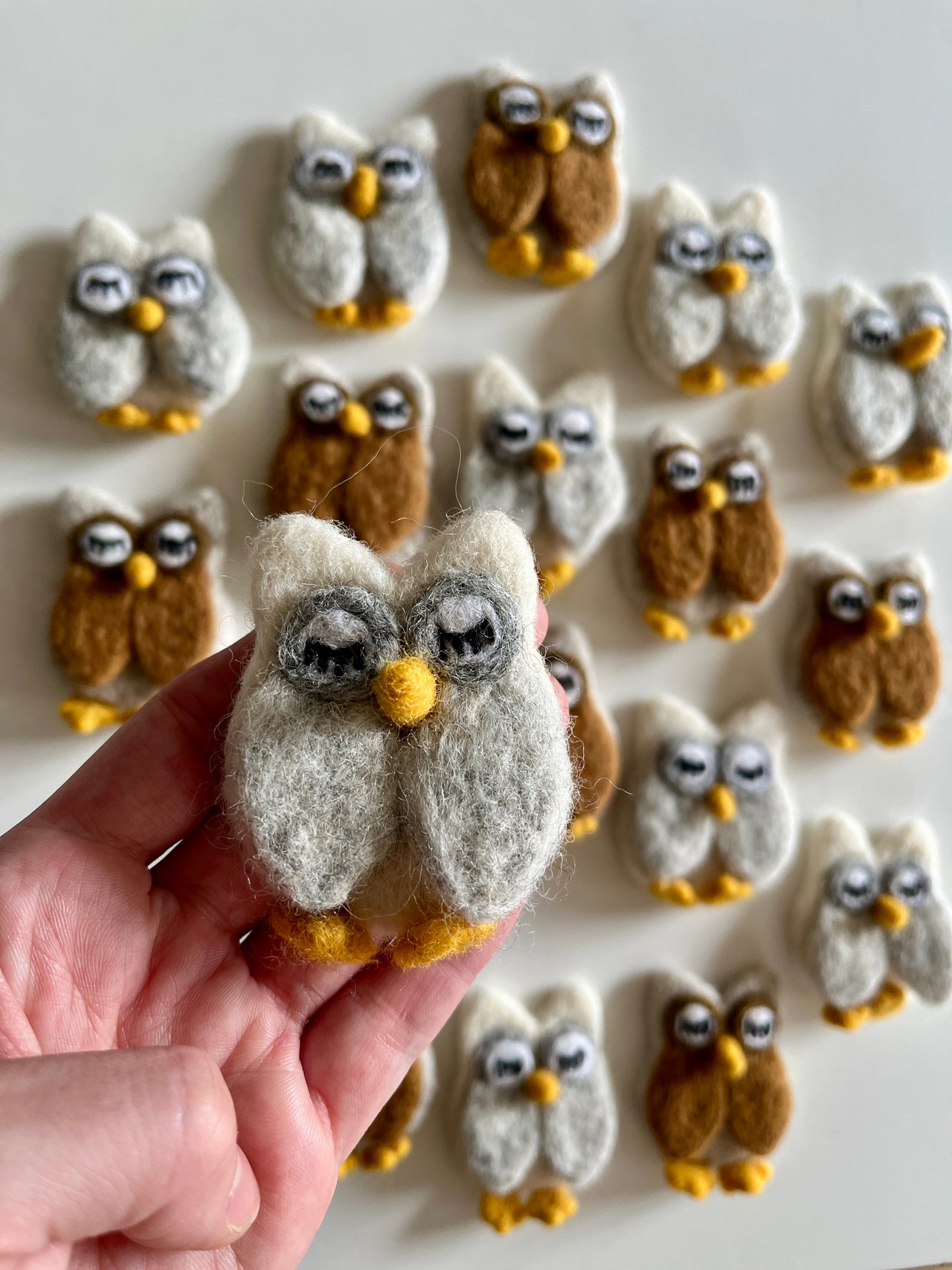 Hand holding a small felt owl figure in gray and white with more similar brown and white felt owls arranged on white surface behind