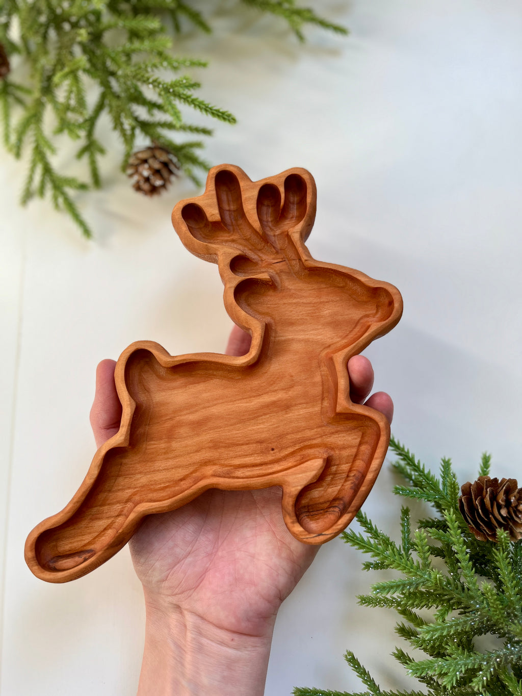 Hand holding a carved wooden tray shaped like a leaping reindeer with pine branches and pine cones nearby on white surface