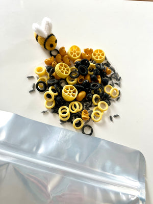 Black and yellow pasta shapes mixed with black rice grains beside a small felt bee and a partially visible silver bag