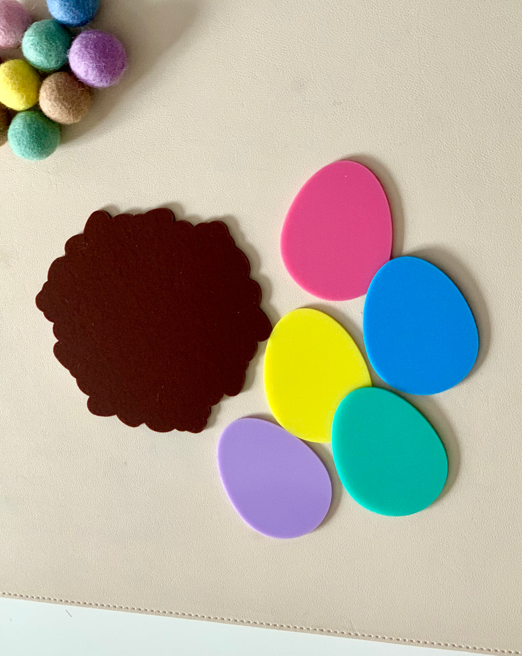 Brown scalloped felt piece beside colorful egg-shaped cutouts and pastel pom-poms on a beige surface