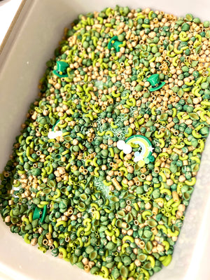 Container filled with green and beige pasta shapes, small beads, and a rainbow and green hat charms scattered inside